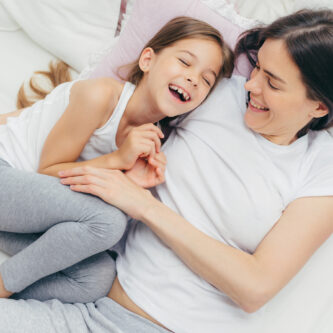 Photo of adorable small child and her mother have fun together in bed, tickle each other, smile joyfully, play after good sleep, have good relationships. People, family, motherhood and childhood